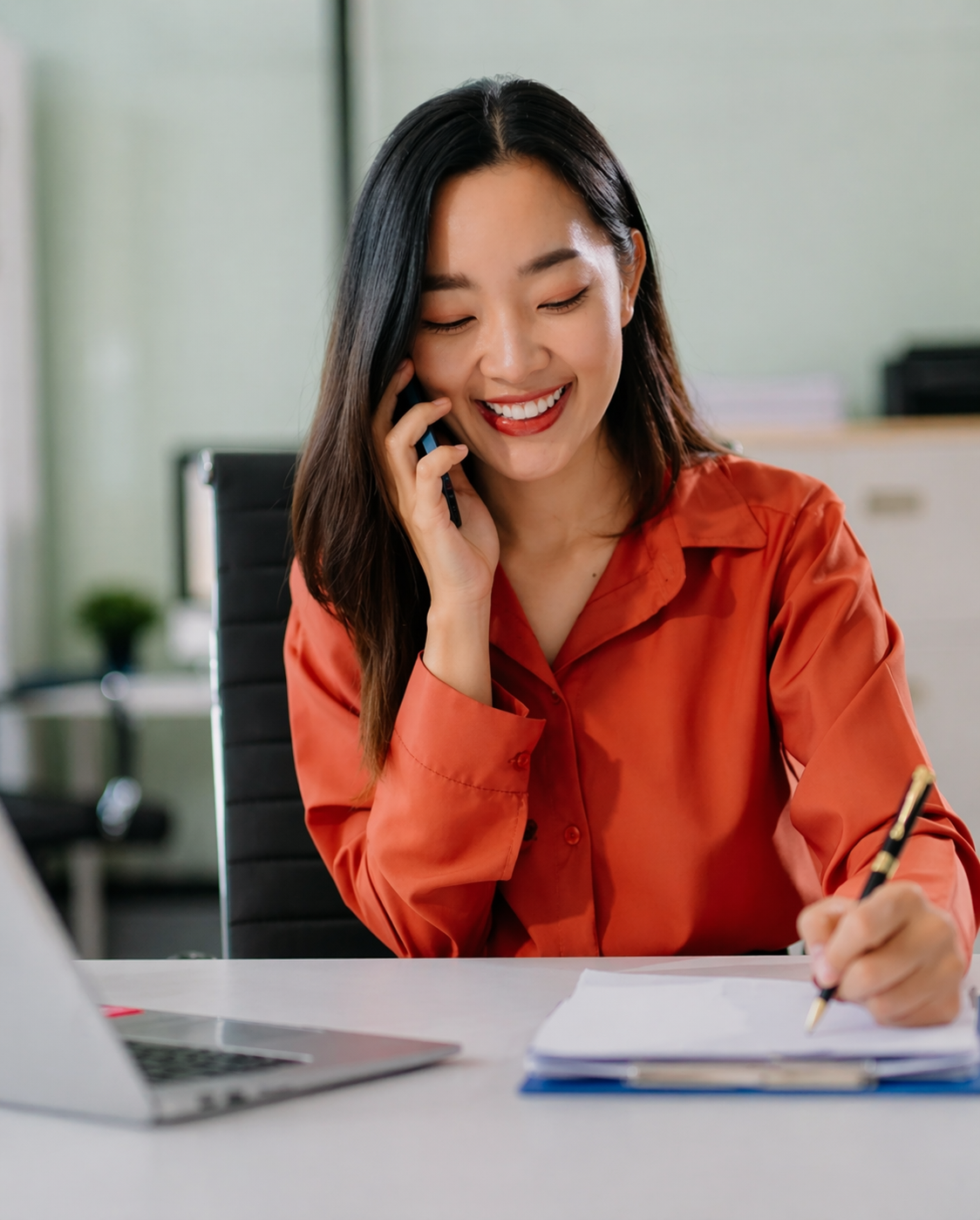 Smiling woman talking on the phone while writing notes at her desk